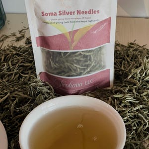 May include: A white ceramic teacup filled with light brown tea, next to a bag of Soma Silver Needles. The bag has text that reads "Divine nectar From Himalayas Of Nepal" and "Handpicked young buds from the Nepal highlands."