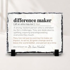 May include: A rectangular white stone plaque with a rough, dark grey chiseled edge, supported by two black plastic stands. The plaque features black and brown printed text defining the term difference maker and includes the name Dr. Sean Hendricks.