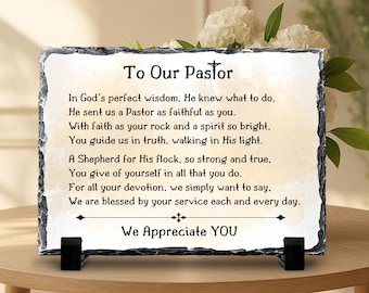 May include: A rectangular slate plaque with rough, chiseled edges, featuring a printed poem titled To Our Pastor with a cross symbol. The text expresses gratitude and is supported by two black plastic stands on a flat surface.