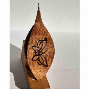 Op de afbeelding: Houten blad-vormige flesstopper met een zwart vlinderontwerp. De stopper is gemaakt van gepolijst hout en heeft een puntige top. Het wordt weergegeven op een kleine houten standaard.