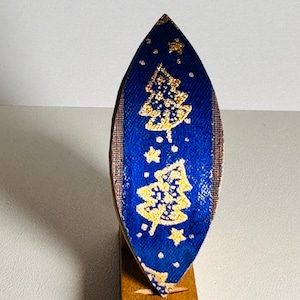 May include: A blue Christmas tree ornament with gold accents, featuring two gold Christmas trees and stars. The ornament is mounted on a small wooden stand, creating a festive holiday decoration.
