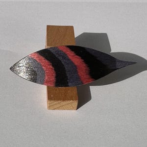 May include: A decorative leaf-shaped object with a striped pattern in black, red, and gray, mounted on a small wooden stand. The leaf has a pointed tip and a shiny surface, creating a contrast with the matte wood.