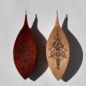 Op de afbeelding: Twee houten oorbellen in een druppelvorm. De ene is donkerroodbruin gebeitst, de andere heeft een lichte, natuurlijke houtkleur. Beide oorbellen hebben een zwart bloemmotief. De oorbellen hangen op een wit oppervlak.