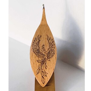 May include: Wooden knitting needle with a phoenix design burned into the wood. The needle is pointed at the top and has a small loop at the end.