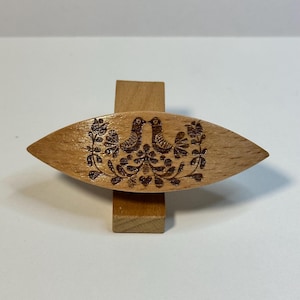 Op de afbeelding: Een houten naaldhouder met een gesneden ontwerp van twee vogels die elkaar aankijken, omringd door bloemen en bladeren. De naaldhouder is lichtbruin van kleur en heeft een gladde, gepolijste afwerking.