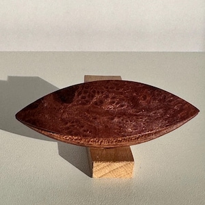 Könnte beinhalten: Eine kleine, bootförmige Schale aus Holz mit einer rotbraunen Oberfläche. Die Schale ruht auf einem kleinen Holzblock.