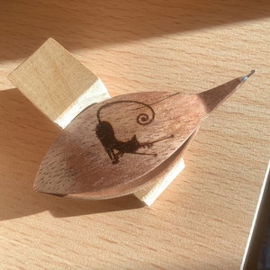 May include: A wooden cat-shaped needle threader with a small wooden stand. The cat is curled up with its tail in the air.