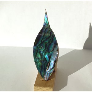 May include: A teardrop-shaped ornament made of iridescent abalone shell with a green, blue, and purple color scheme. The ornament is mounted on a wooden stand.