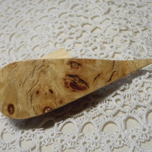 Op de afbeelding: Een handgemaakte haaknaald van lichtgekleurd hout met een uniek nerfpatroon. De haaknaald heeft een druppelvormige handgreep en een zilveren metalen haak aan de punt. De houten handgreep is ongeveer 15 cm lang.