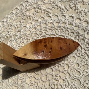 Op de afbeelding: Een houten bobbin-kant shuttle met een natuurlijke houten afwerking. De shuttle is gemaakt van twee stukken hout die aan elkaar zijn bevestigd. De shuttle ligt op een witte kantdoily.