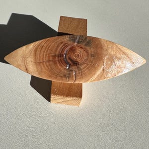 May include: A wooden oval-shaped object with a natural wood grain pattern and a circular knot in the center. The object is resting on a small wooden block.