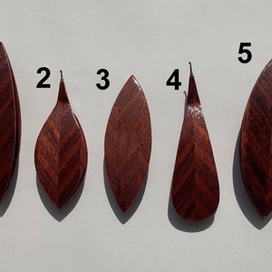 Op de afbeelding: Vijf houten hangers genummerd van 1 tot 5, elk met een gepolijste, roodbruine houtnerf met een duidelijk visgraatpatroon. De vormen variëren tussen langwerpige bladvormen en druppelvormige silhouetten, allemaal met een gladde, glanzende afwerking.