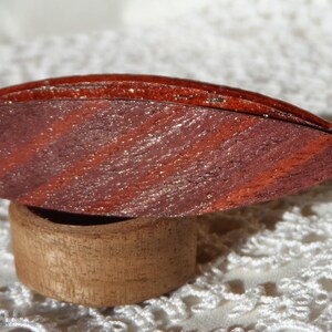 May include: A wooden ring box with a red and brown wood grain pattern. The box is open and the lid is resting on top of the box.