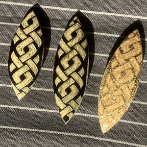 May include: Three leaf-shaped decorative ornaments with a woven design. Two are black with gold-colored accents, and one is gold with a gold-colored woven design. The ornaments are displayed on a striped background.