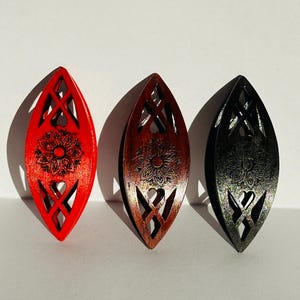 Op de afbeelding: Drie langwerpige, puntige, ovaalvormige houten haaraccessoires met ingewikkelde lasergesneden rasterpatronen en een centrale bloemengravure. De set bevat een felrood, een donkerbruin en een zwart exemplaar, elk met een gladde afwerking.