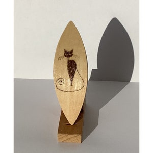 May include: A wooden cat-shaped decorative item. The light-colored wood has a dark brown cat illustration with a glittery body. The cat is standing on a small wooden base. The item casts a shadow on the white background.
