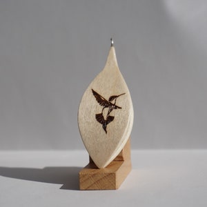 Op de afbeelding: Een lichtgekleurde houten hanger in de vorm van een blad, met een gedetailleerd ontwerp van een kolibrie. De kolibrie is afgebeeld met uitgespreide vleugels en is geëtst in een donkerbruine kleur. De hanger hangt aan een kleine zilveren lus en staat op een houten standaard.