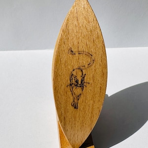 Op de afbeelding: Een lichtbruin houten weefspoel met een puntig, amandelvormig profiel. Het oppervlak is voorzien van een donkere, fijn getekende pyrografie-illustratie van een kat die naar voren loopt met de staart omhoog, gecentreerd op de houtnerf.