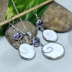 Puede incluir: Un conjunto de collar y pendientes de plata con piedras preciosas ovaladas moradas y colgantes de concha blanca con diseños en espiral. Las joyas se exhiben sobre una superficie de piedra.