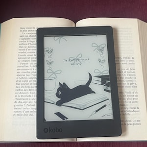 May include: A black Kobo e-reader displaying a cat illustration and the text "my own personal library" rests on an open book. The e-reader is centered, with the book's pages visible on either side. The background is a dark red surface.