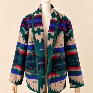 May include: A long-sleeved open-front jacket featuring a geometric pattern in shades of tan, dark green, royal blue, and red. The garment has a shawl collar, two front patch pockets, and dark green trim along the edges and pocket openings.