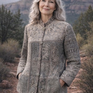 May include: A woman wearing a textured, button-down jacket with a patchwork design in shades of gray and beige. The jacket has a mandarin collar and pockets. The background features a mountain landscape and a dirt path.