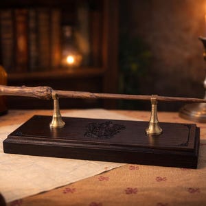 Op de afbeelding: Een houten toverstaf met een getextureerde handgreep rust op een donkerbruine rechthoekige basis met messing steunen. De basis heeft een gegraveerd ontwerp. De toverstaf wordt tentoongesteld op een tafel met een kaars en boeken op de achtergrond.