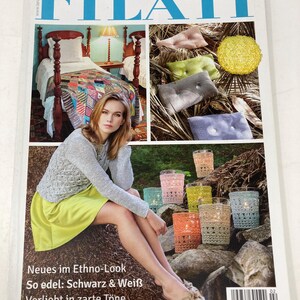 May include: A magazine cover featuring a woman in a gray sweater and yellow skirt. The cover includes images of a bed with a quilt, decorative pillows, and colorful candle holders. Text on the cover reads "home FILATI" and "Neues im Ethno-Look".
