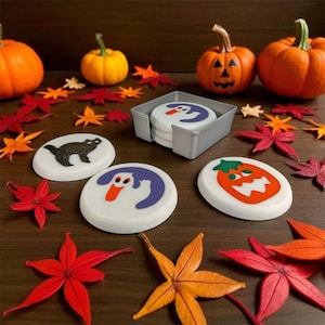 May include: A set of Halloween-themed coasters with a gray holder. The coasters feature various designs, including a black cat, a ghost, and a jack-o'-lantern. The scene is decorated with orange pumpkins and colorful fall leaves, creating a festive autumn display.