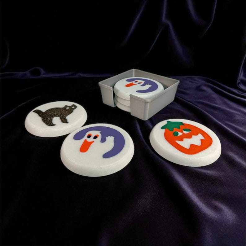 May include: A set of Halloween-themed coasters with a gray holder. The coasters feature white backgrounds with colorful illustrations of a black cat, a ghost, and a jack-o'-lantern. The coasters are round and appear to be made of a durable material.