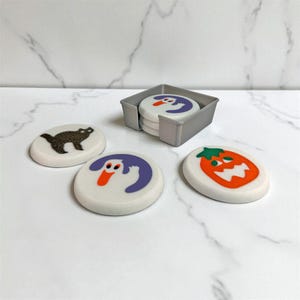 May include: A set of Halloween-themed coasters with a gray holder. The coasters feature a black cat, a ghost, a pumpkin, and a ghost design. The coasters are white with colorful illustrations.