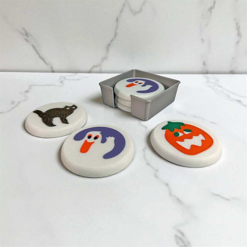 May include: A set of Halloween-themed coasters with a gray holder. The coasters feature a black cat, a ghost, a pumpkin, and a ghost design. The coasters are white with colorful illustrations.