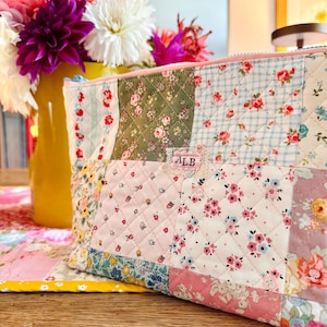 May include: A quilted zipper pouch featuring a patchwork design of various floral and gingham fabrics in shades of pink, green, blue, and white. A small rectangular fabric label with the letters ALB is stitched to the center of the pouch.