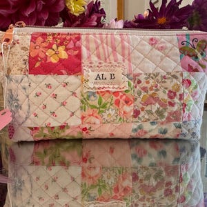 Peut inclure: Une pochette zippée en patchwork matelassé composée de divers carrés de tissu à motifs floraux et rayés dans des tons de rose, blanc et vert. Une petite étiquette rectangulaire avec le texte AL B est cousue au centre, et un motif papillon est visible.