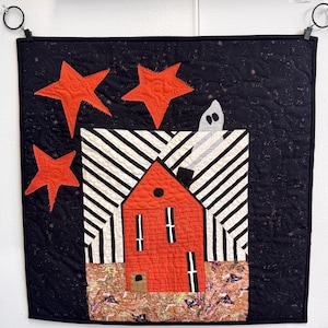 May include: A square fabric wall hanging featuring a red house with black and white striped roof sections, a white ghost emerging from the chimney, and three orange stars against a dark black background with a textured, mottled fabric base.