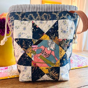 May include: A quilted drawstring project bag featuring a central star block pattern with a graphic illustration of a person knitting. The fabric displays navy blue, cream, and yellow floral prints, a grey striped interior lining, and a brown leather handle.