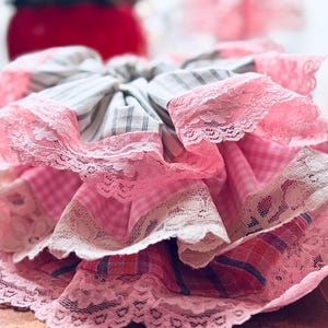 May include: A layered scrunchie featuring pink gingham, striped, and plaid fabrics. The scrunchie is trimmed with delicate pink lace, creating a feminine and vintage aesthetic. The scrunchie is a hair accessory.