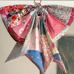 May include: A large decorative bow constructed from various quilted fabric patches featuring floral patterns, gingham checks, and solid colours in shades of pink, red, blue, and white. The bow is suspended from a silver metal ring at the top centre.