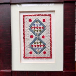 May include: Framed textile art piece featuring a central design of two connected diamond shapes with a patchwork pattern in red, blue, and white. Red star accents and a red border surround the design. The frame is a dark, reddish-brown color.