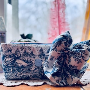 May include: A blue and white toile-patterned cosmetic bag with a white lace trim and a matching bow. The bag and bow are on a lace doily, with a window in the background.
