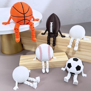 Op de afbeelding: Een verzameling sportbalfiguren, waaronder een basketbal, een voetbal, een honkbal, een volleybal, een golfbal en een voetbal. Elke bal heeft beweegbare benen en armen en zit of rust op een oppervlak. De basketbal is oranje, de voetbal is bruin en de honkbal is wit met rode stiksels.