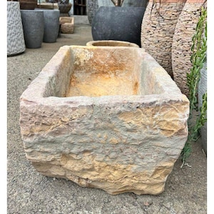 May include: A rectangular stone planter with rough, textured sides. The planter has a natural color palette of tan, gray, and brown. The open top reveals the interior. Other planters are visible in the background.