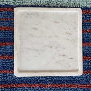 May include: A square, white marble tray with a slightly raised edge. The marble has subtle gray and brown veining. The tray is set against a blue, red, and green striped background.