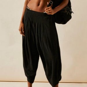 Puede incluir: Pantalones harem negros con cintura alta y corte holgado. Los pantalones se combinan con sandalias de plataforma negras y un top de bikini estampado. Una mochila negra se lleva sobre el hombro. El conjunto es informal y cómodo.