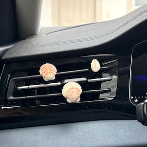 May include: Three seashells, in shades of white and orange, are displayed on a car's air vent. The shells are arranged in a decorative manner, adding a coastal touch to the vehicle's interior. The car's dashboard is black and gray.