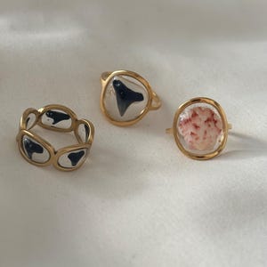 May include: Three gold-toned rings. One ring features a black shark tooth set in clear resin. Another ring has a white and red seashell. The third ring has a series of black shark tooth shapes set in clear resin.