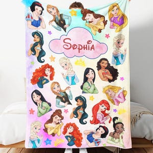 May include: A personalized blanket featuring cartoon illustrations of various princesses. The blanket is predominantly white with a colorful design, including stars and a cloud with the name "Sophia" in pink script. The princesses are depicted in their signature outfits.