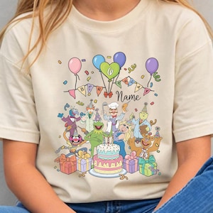 May include: A beige t-shirt featuring a colorful graphic of Muppet characters celebrating a birthday. The design includes a cake, balloons, and the word "Name". The shirt is worn by a person wearing blue jeans.