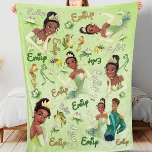 May include: A light green blanket featuring illustrations of a princess in a green dress, a frog, and the name "Emily" in gold script. The blanket has a repeating pattern of the princess and other elements.
