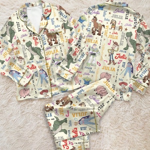 Puede incluir: Un conjunto de pijama de camisa de manga larga con botones y pantalón a juego en color crema, con un patrón repetitivo de personajes de Toy Story, estrellas y el nombre Julia impreso en varios colores y tipografías sobre la tela.